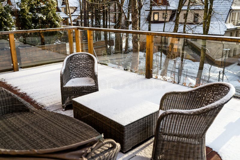Wicker garden furniture on a wooden, snowy terrace with glass balustrades, in the background trees and houses. royalty free stock photo